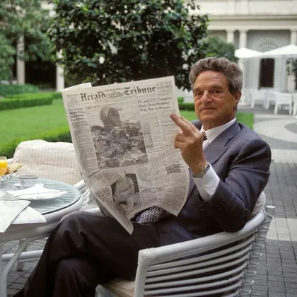George Soros, in a grey suit, holding up a copy of the International Herald Tribune while seated at an outdoor breakfast table